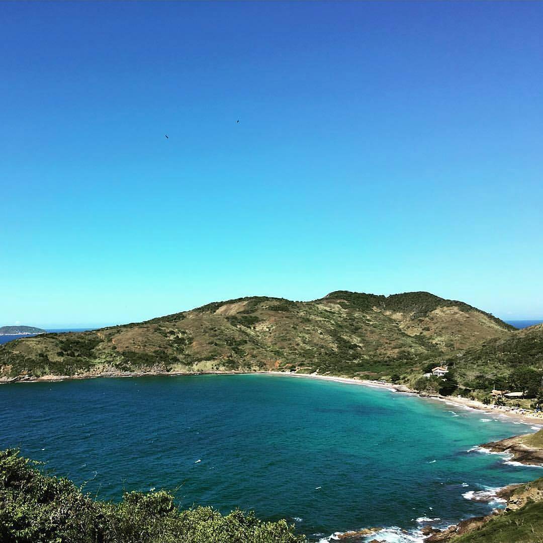 Uma vista dramática das ondas batendo nas rochas da Praia Brava em Búzios, com as falésias verdes da costa ao fundo.